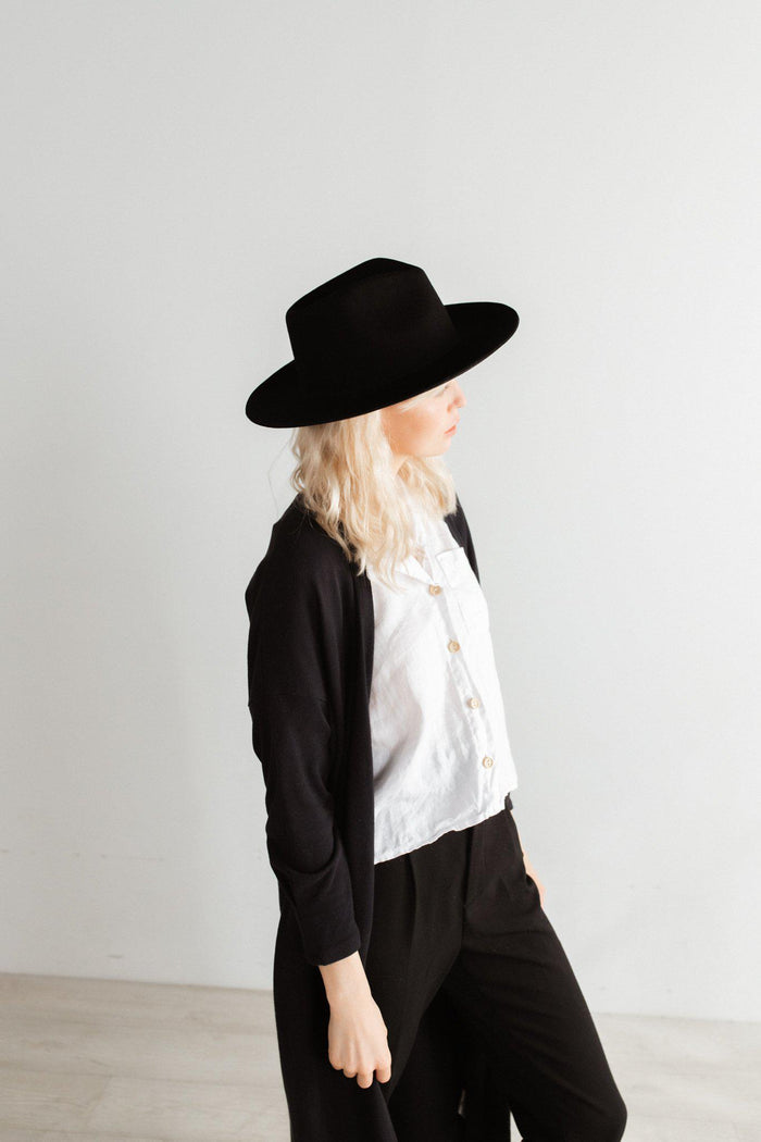 Woman wearing a black fedora hat, a white shirt with a black coat and black pants, looking sideways against a plain background #color_black
