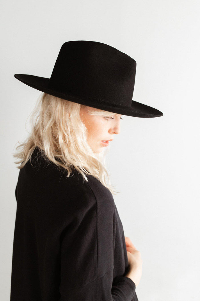 Woman wearing a black fedora hat and a black coat, facing sideways against a plain background #color_black