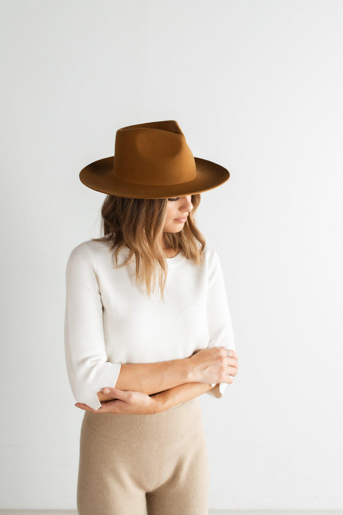 Woman wearing a cinnamon colored fedora hat, a white shirt and beige pants against a plain background #color_cinnamon