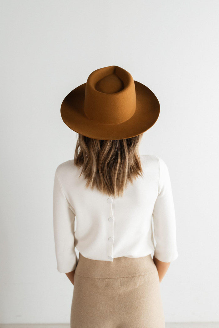 Woman wearing a cinnamon colored fedora hat, a white shirt and beige pants, facing behind against a plain background #color_cinnamon