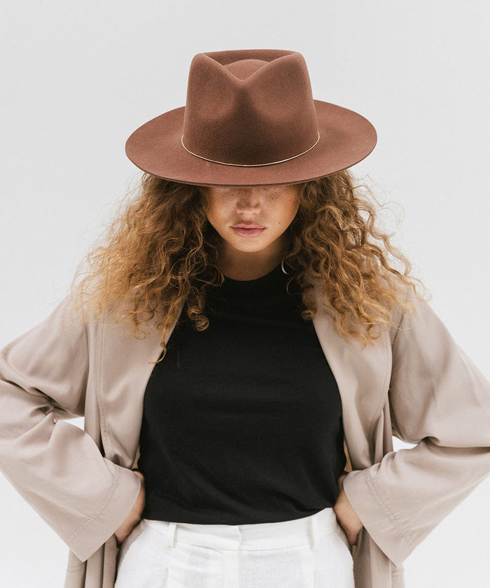 Woman wearing a dark oak colored fedora hat with a hat band, a black shirt and a beige jacket against a white background #color_dark oak