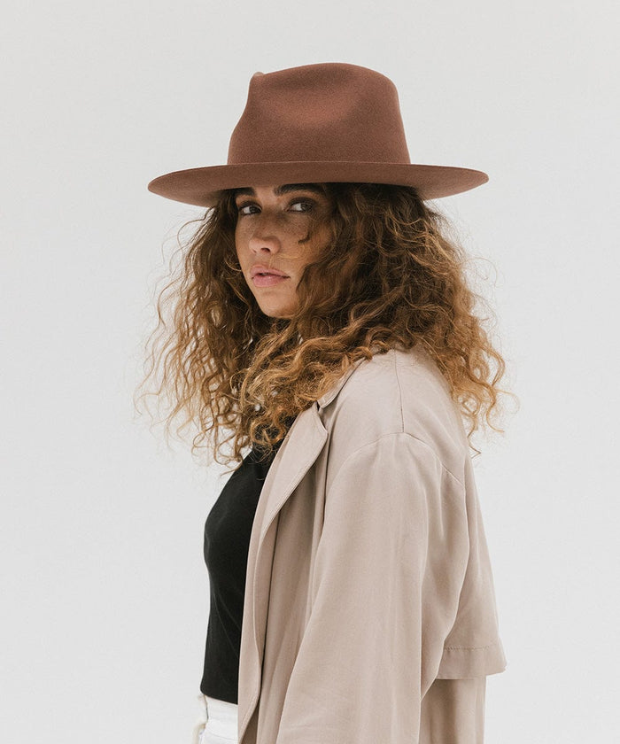 Woman wearing a dark oak colored fedora hat and a beige jacket, posing sideways against a white background #color_dark oak