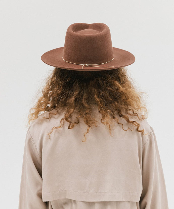 Woman wearing a dark oak colored fedora hat with a hat band and a beige jacket, facing behind against a white background #color_dark oak