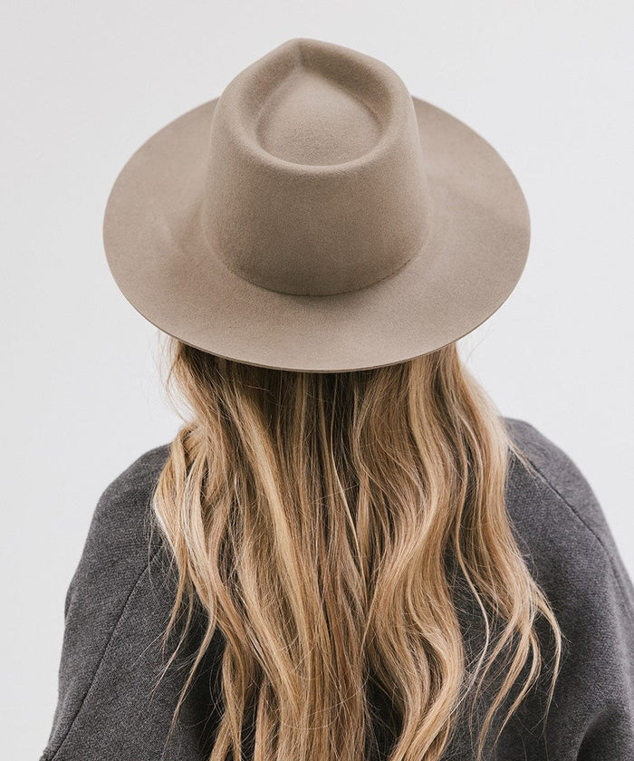 Woman wearing a sage fedora hat and a dark grey jacket, facing behind against a white background #color_sage